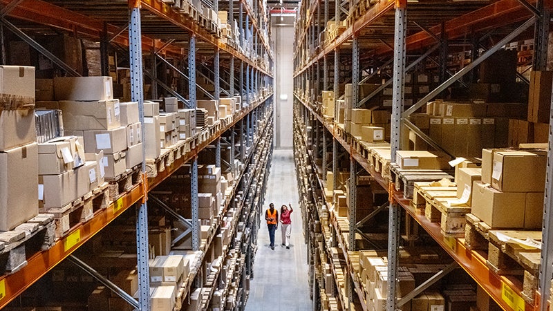 Two manufacturing workers walk down warehouse aisle surrounded by tall industrial shelves.